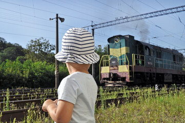 Child in striped hat watching a passing locomotive on sunny summer day. Railway, childhood curiosity and travel theme.
