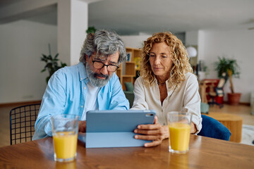 Senior couple using digital tablet and drinking orange juice while sitting at dining table in their modern apartment