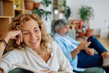 Smiling mature woman enjoying a book while relaxing at home, her husband is using a smartphone in the background