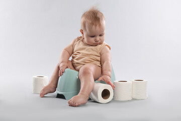 Cute little baby sitting on potty and rolls of toilet paper against light grey background