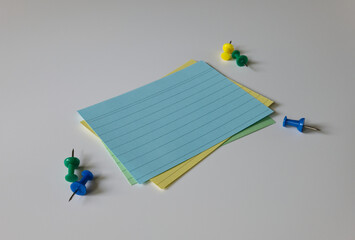 Side view of blank index cards and push pins on a white background. A stack of colorful empty note...