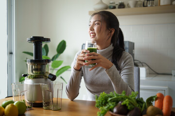 Smiling woman relaxing with a glass of freshly prepared cold-pressed green juice in a home kitchen....