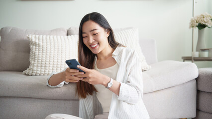 Happy young asian woman sitting on floor and using smartphone at home.