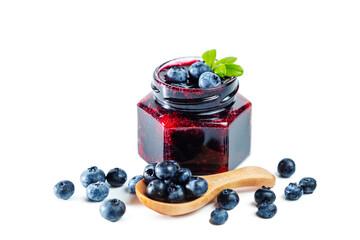 Jar of blueberry jam with berries, leaves and wooden spoon isolated on white background.