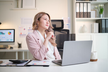 Professional businesswoman looking thoughtful while working on a laptop with financial reports on...
