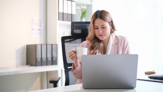Professional businesswoman relaxing with a coffee cup while continuing work on her laptop at desk.