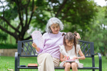 Elderly woman fans herself while watching her granddaughter draw on a park bench.