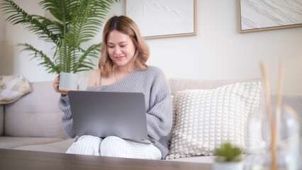 Happy female freelancer enjoying remote work with laptop and hot beverage in modern living room.