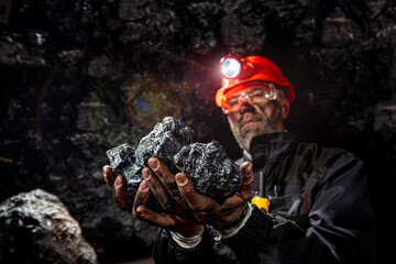 Coal miner with a dirty face in black dust, wearing a protective helmet with a flashlight and work...