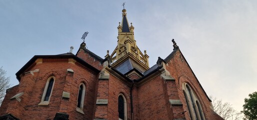 A low-angle shot of a red brick church with a towering central spire. The ornate architecture features arched windows, and crosses adorn the roofline. The sky above is a clear, pale blue.