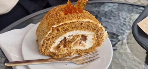 A delicious slice of honey cake on a white plate, it's a rolled cake with cream and caramel filling, topped with crumbs and a piece of honey candy
