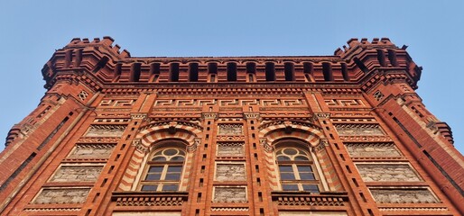 A grand red brick building with an ornate facade stands against a clear blue sky. The architecture features classical Greek patterns, twin arched windows, 