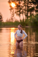 Fototapeta premium Young woman with long dark hair standing in calm lake during sunset. Golden light reflects on water, surrounded by lush trees. Peaceful atmosphere with warm tones