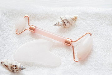 Rose quartz facial roller and gua sha stone arranged on a soft white towel with decorative seashells for a skincare and beauty spa concept