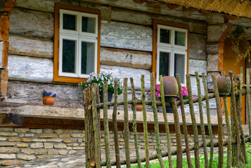 Old wooden cottage with whitewashed windows and flowers, rustic branch fence with clay pots in...