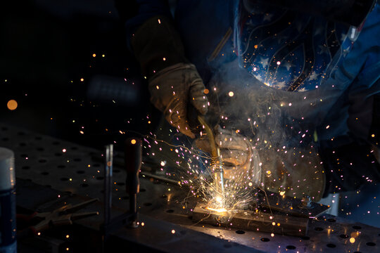 Man with welding mask in safety suit with gloves welds steel in home workshop. Welding flame with sparks. Dangerous work
