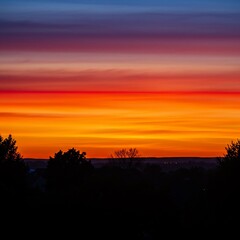 Fototapeta premium Vibrant Sunset Sky with Layered Colors of Orange Red Yellow Blue and Silhouetted Trees Against the Horizon During Dusk