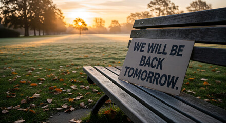 Hopeful 'WE WILL BE BACK TOMORROW' sign on park bench at sunrise in a misty autumn park