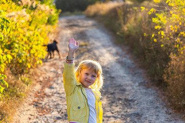 Young girl waving goodbye on a sunny trail with a dog in the background during autumn