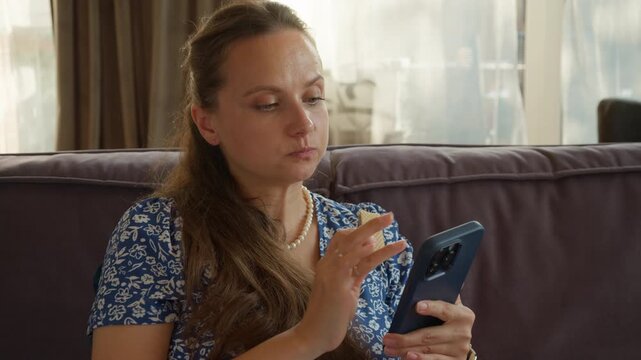 A girl eats a sugary wafer, a processed junk food with high calories. An example of an unhealthy diet, bad eating habits, and mindless consumption while being distracted by her smartphone at home.