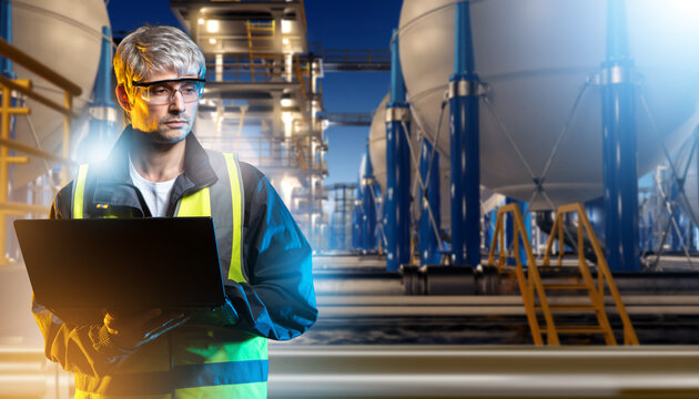 Man refinery engineer. Specialist with laptop near high pressure tanks. Man inspector servicing BPVC with cryogenic liquids. Refinery contractor stands under night sky. Industrialist quarry