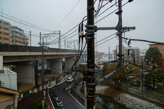 Snow falls over tangled power lines and city streets, blending the industrial with the seasonal in a quiet urban winter scene.