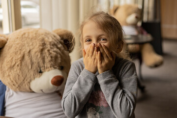 A happy young girl covers her mouth while smiling, standing near a large stuffed teddy bear in a...