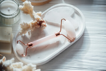Rose quartz face roller and gua sha stone on a white ceramic tray with coral decor and a lit candle...