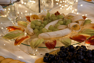 Festive Fruit Platter Arrangement with Bananas, Grapes, Apples and Cherries Illuminated by Warm String Lights, White Tablecloth Setting