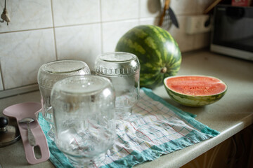 Glass jars prepared for preserving stand upside down on a towel, while watermelon is being cut into slices