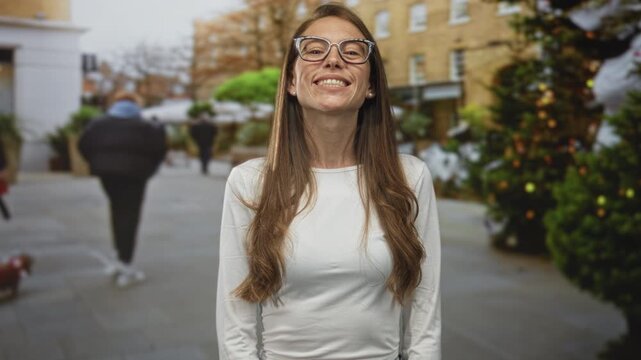 Woman wearing brown framed glasses puckers lips and makes a fish face while standing on a street near a decorated christmas tree; playful curiosity holiday cheer.
