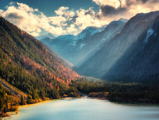 Aerial view of a peaceful alpine lake surrounded by autumn forests and snow-capped mountains at sunset. Sunbeams, illuminating the colorful trees and calm water, sky with clouds in fall. landscape