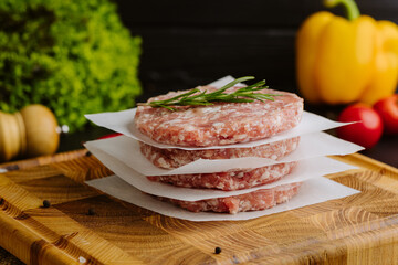 Raw burger patties stacked with parchment and rosemary on a wooden board, ready to cook