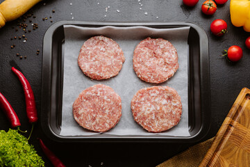 Raw burger patties on baking tray with fresh vegetables and spices on dark background