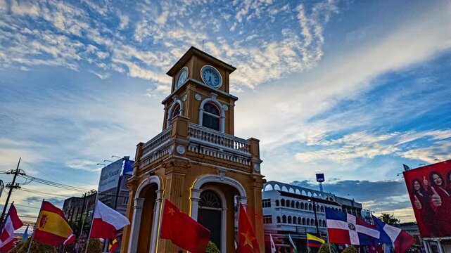 Phuket-Thailand, 13 August 2025: Time lapse A vibrant view of a historic yellow clock tower surrounded by flags and reflected in a pond under a dramatic sunset sky, capturing both cultural charm 