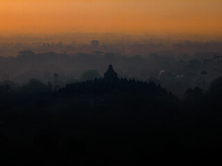 Obraz premium The iconic silhouette of Borobudur Temple emerges from a mystical morning mist, set against the warm, glowing colors of the dawn sky.