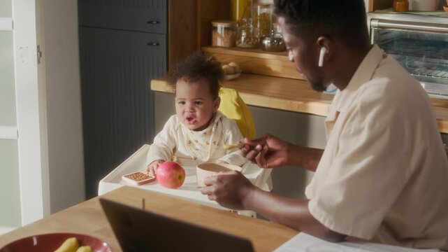 Over shoulder shot of stay at home father feeding cue African American baby sitting in highchair scene lit by sunlight