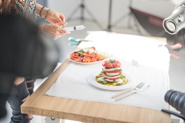 A food stylist, wielding a syringe, meticulously prepares a caprese salad for a photo session. Tomatoes, mozzarella, and basil are arranged artfully on a table
