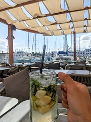 Main tenant un grand verre de mojito frais avec glaçons, citrons et feuilles de menthe, devant un port de plaisance touristique, scène estivale ensoleillée et rafraîchissante, ambiance vacances © HeithemLejmi
