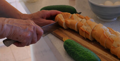 Woman slicing baguette bread at cutting board. Old senior hands and sharp knife.