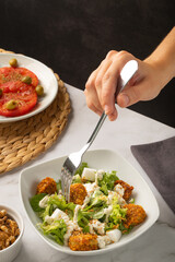 Hand picking with a fork on a caesar salad with vegan nuggets on a white marble table. A side dish of sliced tomato and a small bowl of nuts are sitting next to it. 