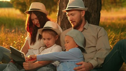 Parents and children play on computer tablet in park under tree. Young family children is sitting on grass in forest relaxing outdoors. Dad mom kids use tablet. Family education with digital gadget - Powered by Adobe