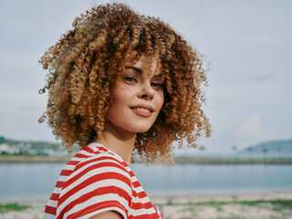 Smiling woman with curly hair wearing a striped red and white shirt outdoors by the water under a...