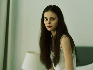 Serious young woman with long hair sitting indoors in soft natural light, wearing a white...