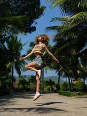 Smiling woman jumps outdoors wearing colorful top and denim shorts under palm trees on sunny summer...