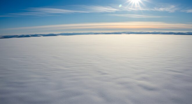 A vast, snow-covered landscape stretches endlessly beneath a bright sun, with a soft layer of clouds floating above the horizon. The serene, expansive view evokes feelings of solitude and infinite bea