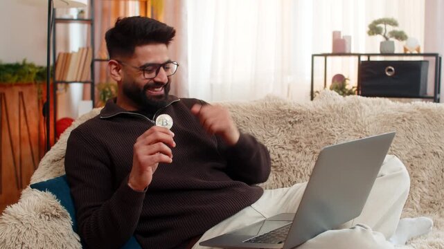 Indian man at home watches Bitcoin BTC chart rise on laptop, smiling while holding coin with confident hand. Arabian guy on sofa shows excitement joy, celebrating crypto success with raised gesture.