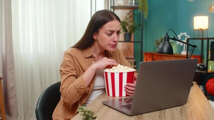 Young woman sitting at home table watches dramatic film on laptop eating popcorn and feeling tense. Caucasian girl grips seat nervously reacting to intense plot twists while chewing popcorn quickly