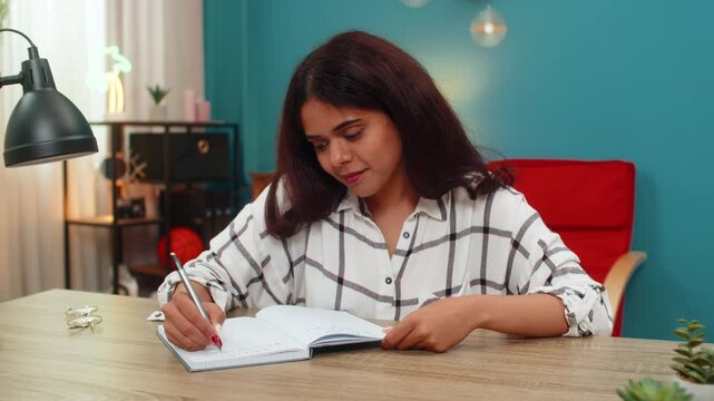 Indian woman at home writes notes in notebook then rereads lines showing careful focus and learning. Arabian girl at table studies written content reviewing details ensuring understanding and mastery.