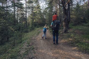 A boy with Down syndrome on his father's shoulders and his sister hiking in the forest. Diverse family walking in nature, together on forest path.